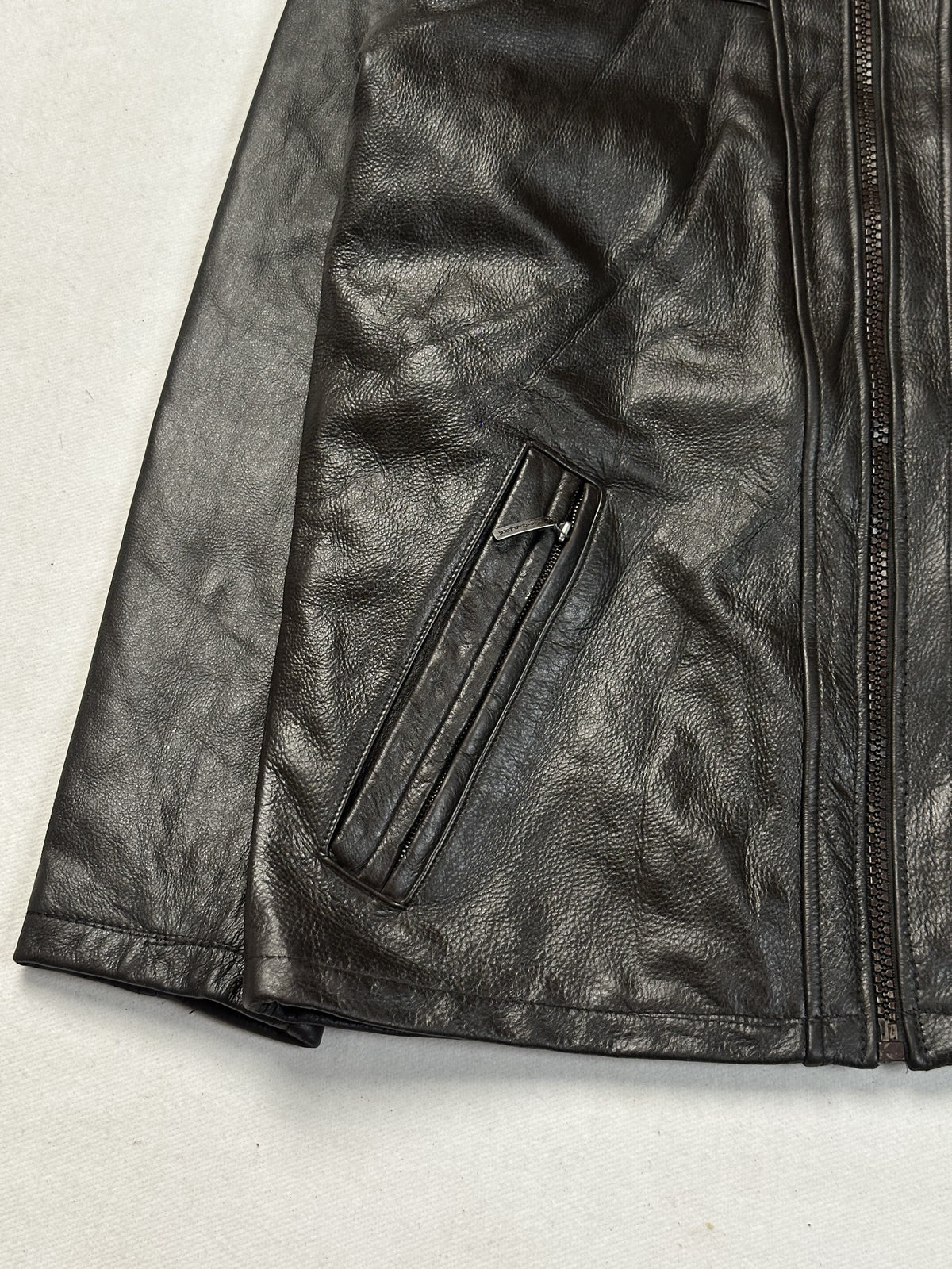 Close view of a diagonal zip pocket and the hem corner on the brown leather jacket