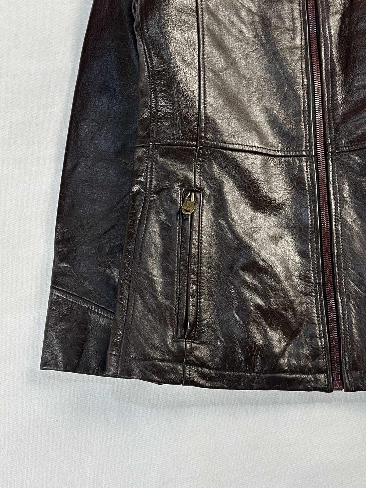 Zipped hand pocket with a brass pull at the hem of the brown leather jacket